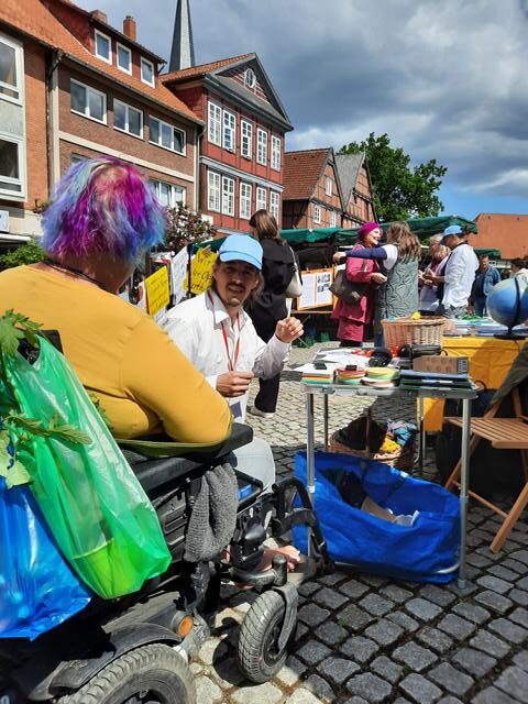 Auf dem Bil ist eine Person mit blauer Schirmmütze zu sehen. Er gehört zu dem Team für die Kunstaktion am 8. Mai. Er unterhält sich mit einer Person im Rollstuhl.