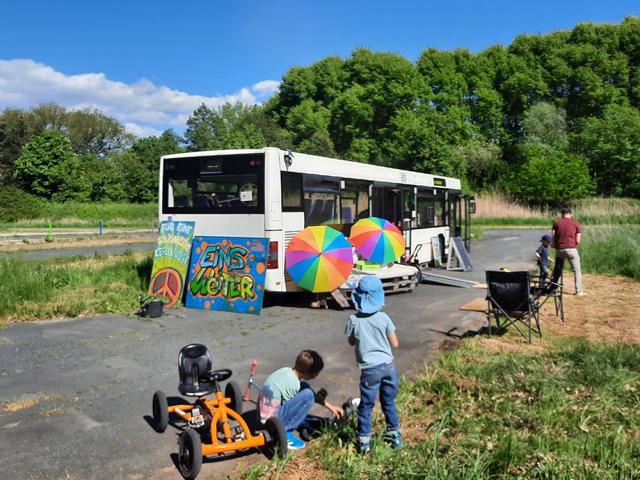 Auf dem Bild ist der alte Bus "Bernhard" zu sehen. Vor dem Bus stehen zwei Palettensofas mit bunten Schirm in Regenbogenfarben darüber. Vorne im Bild sind zwei Kinder von hinten zu sehen, rechts im Bild ein Mann von hinten.
