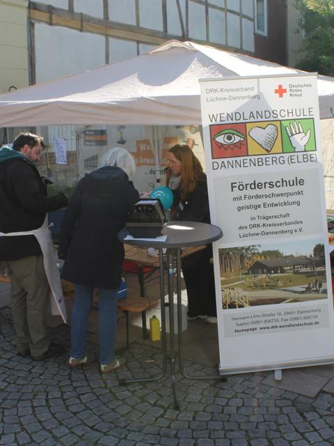 Auf dem Bild sind Menschen von hinten zu sehen, die an einem Hochtisch stehen. Daneben ist ein Informations-Banner von der Wendlandschule in Dannenberg zu sehen.
