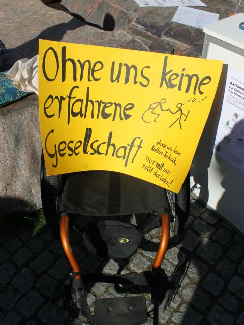 Auf dem Bild ist ein Rollstuhl zu sehen, auf dem ein Plakat plaziert wurde. Auf dem Plakat steht: "Ohne uns keine erfahrene Gesellschaft".