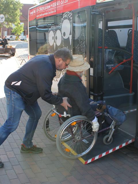 Auf dem Bild ist ein Mann im Rollstuhl zu sehen, der von einem anderen über eine ausgefahrene Rampe in einen Bus geschoben wird.