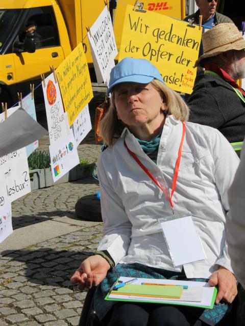 Auf dem Bild ist eine Frau im Rollstuhl zu sehen. Sie trägt eine blaue Schirmmütze auf dem Kopf und hat auf dem Schoß Papiere und Stift liegen. Im Hintergrund sind die Plakate an der Wäscheleine zu sehen.