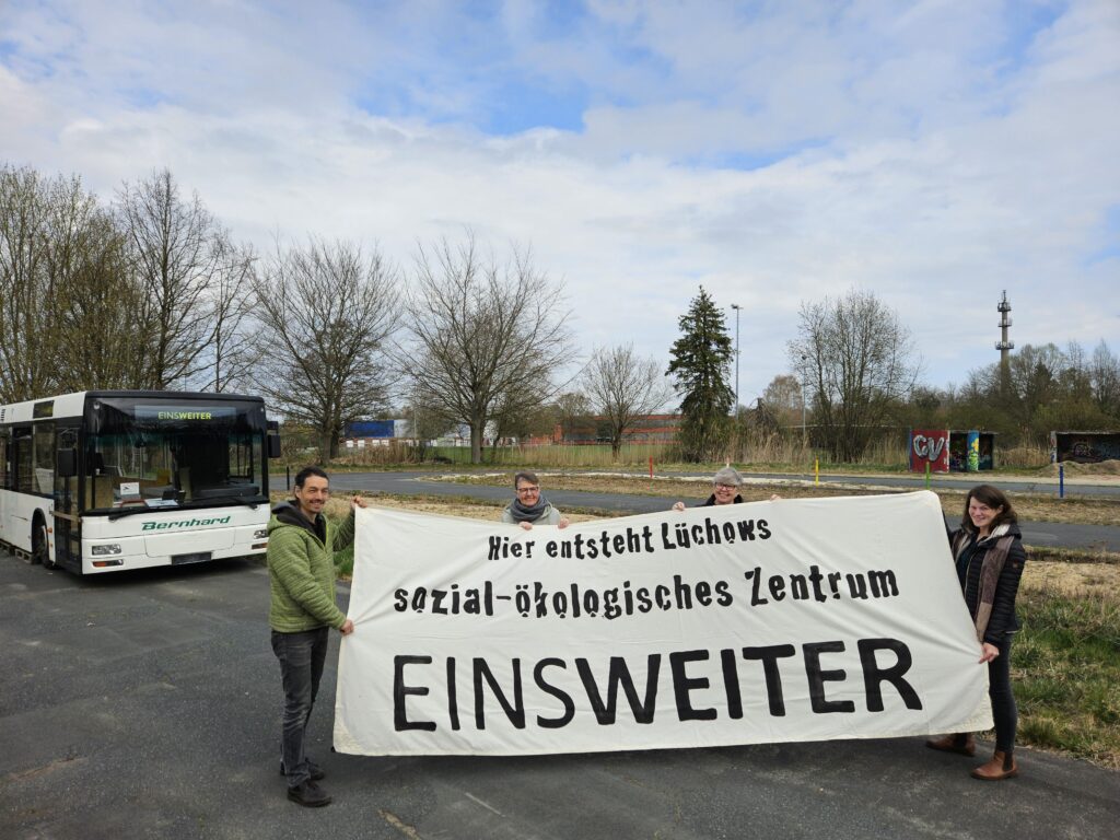 Auf dem Bild sind vier Personen zu sehen, die ein weißes Transparent halten, auf dem mit schwarzer Schrift steht: "Hier entsteht Lüchows sozial-ökologisches Zentrum". Sie stehen auf dem Grundstück von EinsWeiter. Links im Bild ist der alte Bus "Bernhard" zu sehen.