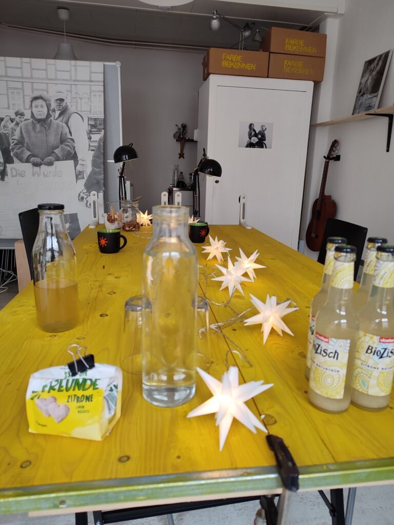 Das Foto zeigt das Büro von EinsWeiter. In der Mitte ist eine gelbe, lange Tischplatte, worauf Getränkeflaschen und Becher stehen sowie eine Lichterkette mit Sternen. Im Hintergrund links hängt ein Plakat mit einer Frau, die ein Schild in der Hand hält mit den Worten: Die Würde des Menschen ist unantastbar. Rechts im Hintergrund steht ein weißer Schrank, an der Wand hängt eine Gitarre.