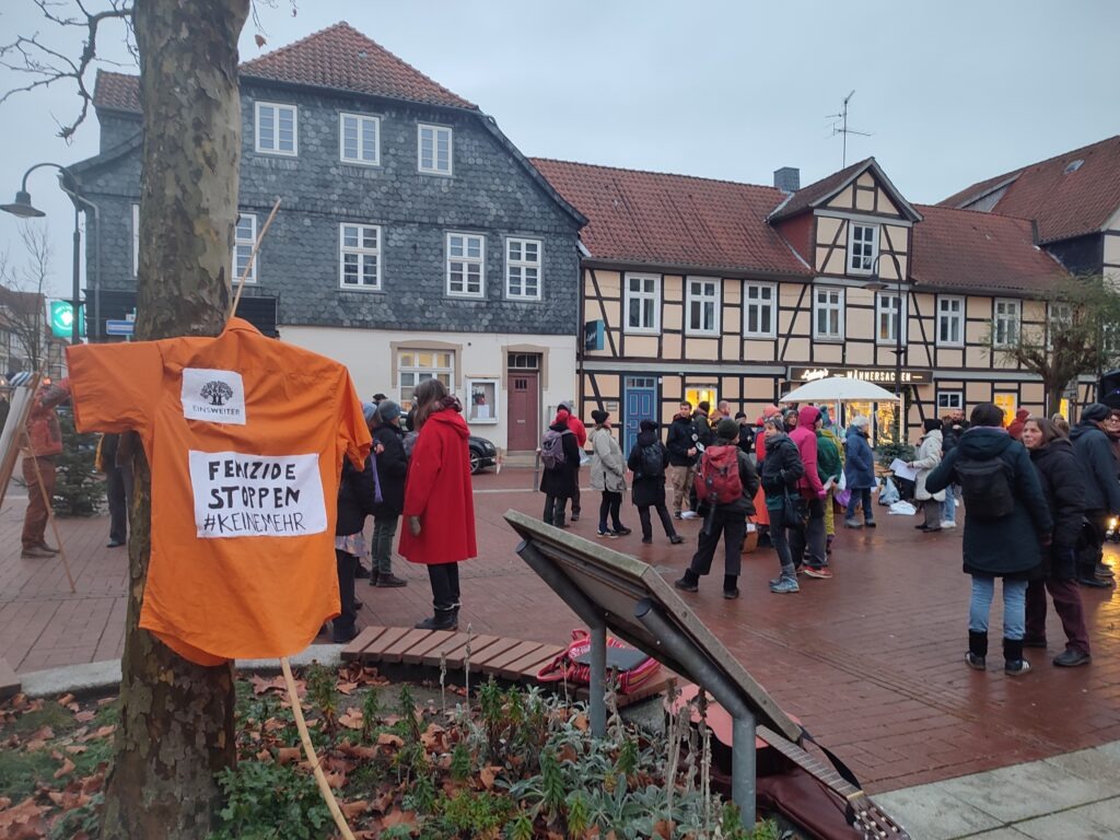 Das Foto zeigt den Marktplatz in Lüchow. Dort stehen auf einer größeren Fläche circa 40 Personen in kleineren Gruppen und unterhalten sich. Ganz vorn hängt in einem Baum ein orangenes T-shirt mit der Aufschrift: Femizide stoppen! Im Hintergrund sind mehrere Fachwerkhäuser, in denen unten teilweise Geschäfte sind.