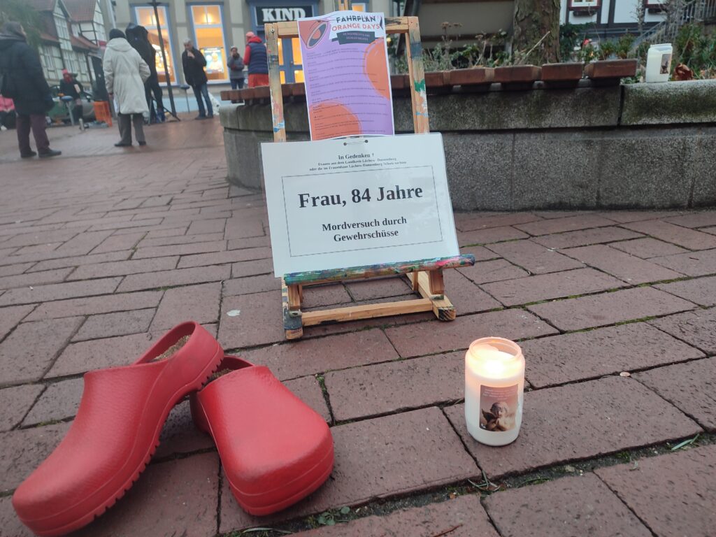 Im Hintergrund ist eine Staffelei mit der Aufschrift: "Frau, 84 Jahre, Mordversuch durch Gewehrschüsse". Rechts steht eine weiße Kerze. Links rote Schuhe. Im Hintergrund sind Menschen, die umher gehen.