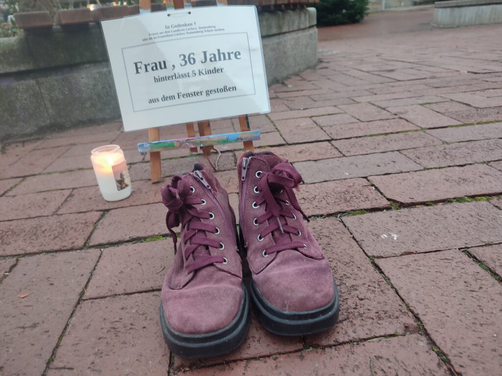 Im Hintergrund ist eine Staffelei mit der Aufschrift: "Frau, 36 Jahre, hinterlässt 5 Kinder, aus dem Fenster gestoßen". Links steht eine weiße Kerze. Vorne rote Schuhe.