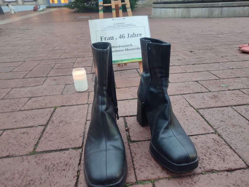 Im Hintergrund ist eine Staffelei mit der Aufschrift: "Frau, 46 Jahre, Mordversuch". Links steht eine weiße Kerze. Vorne schwarze Schuhe.