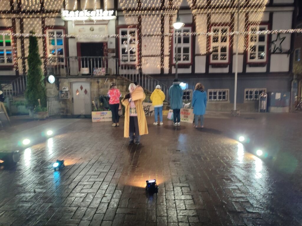 Das Foto zeigt den ausgeleuchteten Markplatz von Lüchow. Hinten sind 5 Menschen von hinten zu sehen. Vorne ist eine Person mit Megaphon.