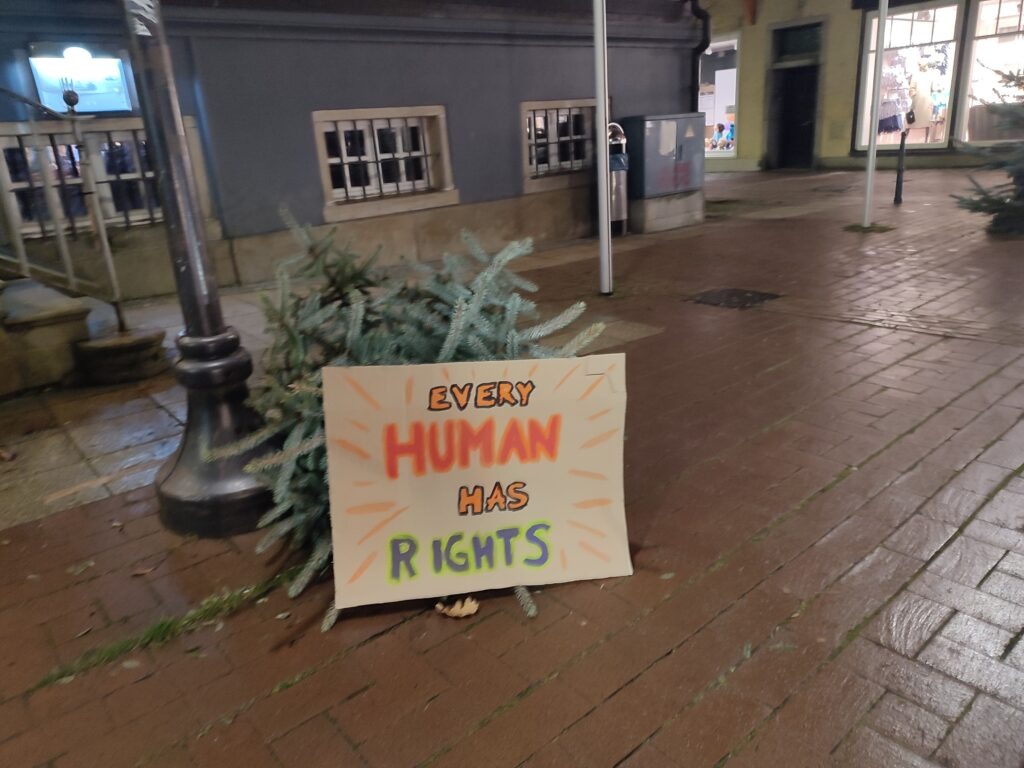 Ein Schild, welches auf dem Markplatz an einem umgefallenen Tannenbaum aufgestellt wurde. In Orange und Grün steht dort: "Every Human has Rights"