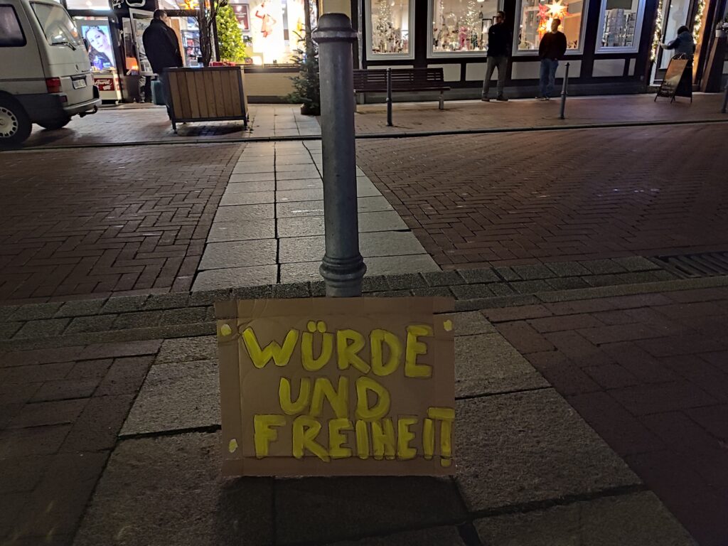 Ein Schild, welches angelehnt an einem Pfahl auf dem Markplatz in Lüchow aufgestellt wurde. Dort steht: Würde und Freiheit. Im Hintergrund ist die Straße und Personen, die in die Schaufenster schauen.
