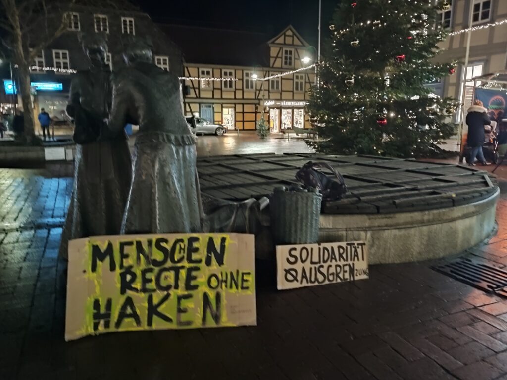 Zwei Schilder, welche auf dem Markplatz in Lüchow aufgestellt wurden. Dort steht: Menschenrechte ohne Haken und Solidarität heißt nicht Ausgrenzung. Die Schilder lehnen an zwei Figuren, die an einem Brunnen stehen, der aber gerade außer Betrieb ist. Ganz im Hintergrund steht ein großer geschmückter Tannenbaum.