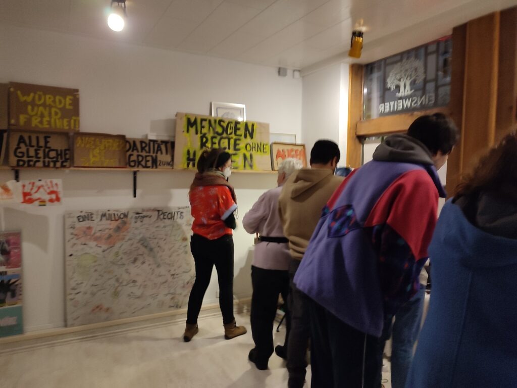 Im Büro von EinsWeiter befinden sich 5 Menschen. Im Hintergrund sind viele verschiedene Schilder, auf denen steht unter anderem:  Würde und Freiheit. Alle Gleiche Rechte. Menschenrechte ohne Haken. 