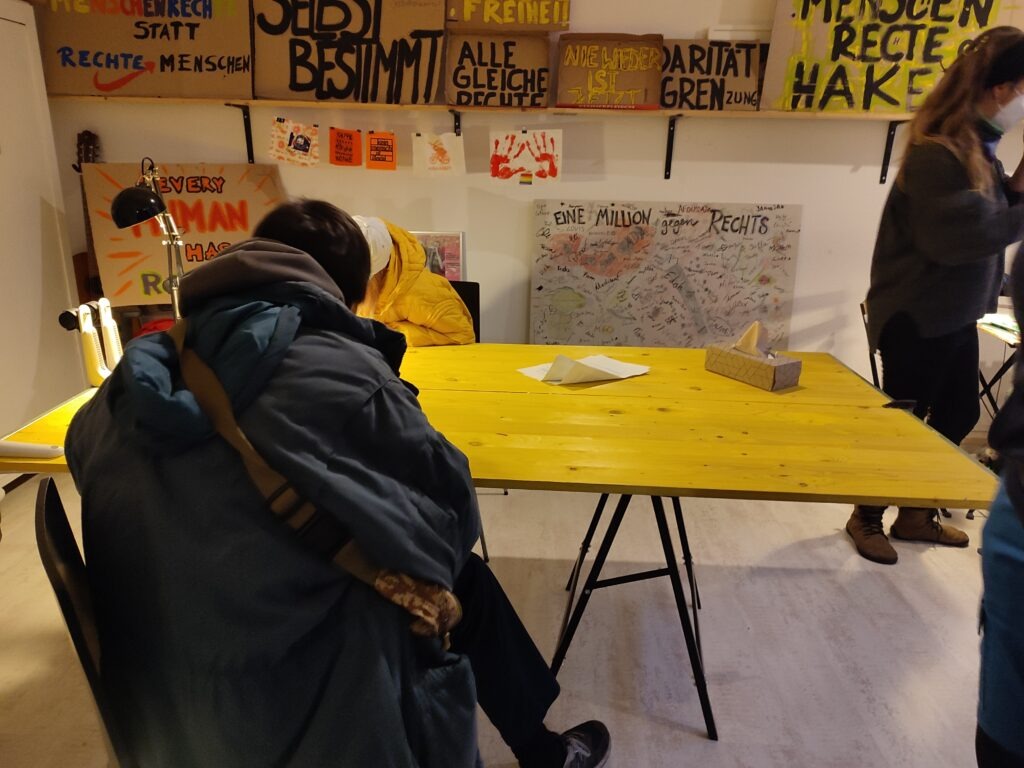 Das Bild zeigt das Büro von EinsWeiter befinden sich zwei Menschen. Im Hintergrund sind viele verschiedene Schilder, auf denen steht unter anderem:  Würde und Freiheit. Alle Gleiche Rechte. Menschenrechte ohne Haken. Im Zentrum ist der gelbe Tisch und eine Person beugt sich über diesen. 