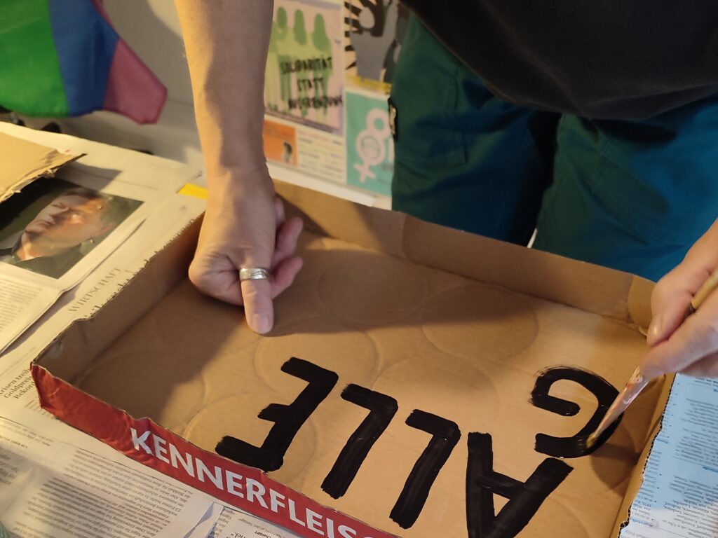 Eine Person malt ein Plakat. Bis jetzt steht da "Alle g..." der Pinsel malt gerade das "g".