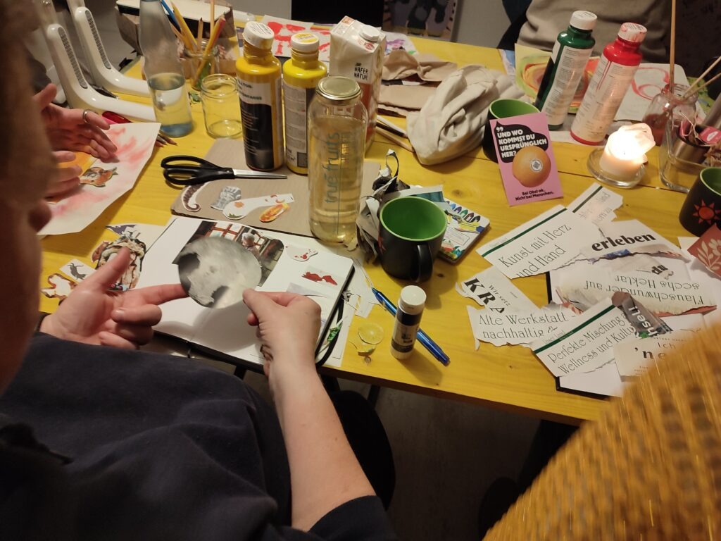 Zentral ist der gelbe Tisch des Büros von EinsWeiter. Auf diesem Tisch sind Zeitungen, Bastelmaterial, Pinsel und Farben. Im Vordergrund ist eine Person, die ein Bild auf ein Plakat klebt. 