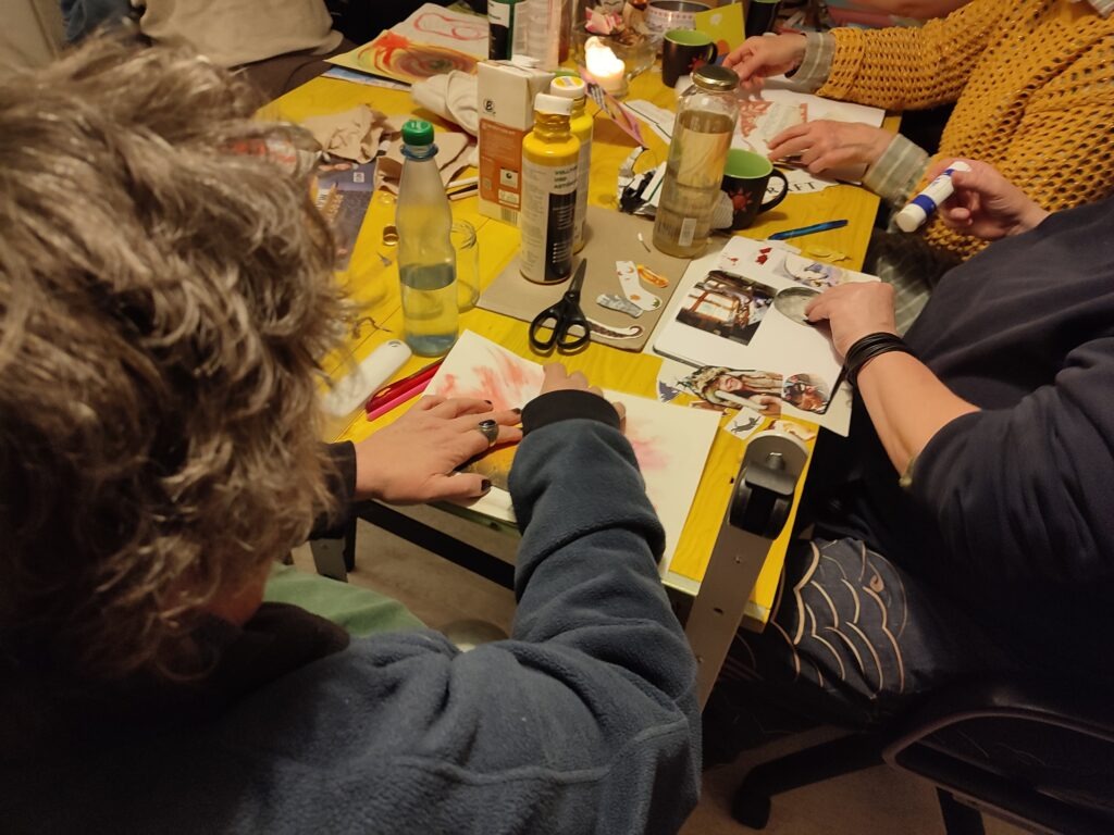 Zentral ist der gelbe Tisch des Büros von EinsWeiter. Auf diesem Tisch sind Zeitungen, Bastelmaterial, Pinsel und Farben. Im Vordergrund ist eine Person, die ein Bild auf ein Plakat klebt.