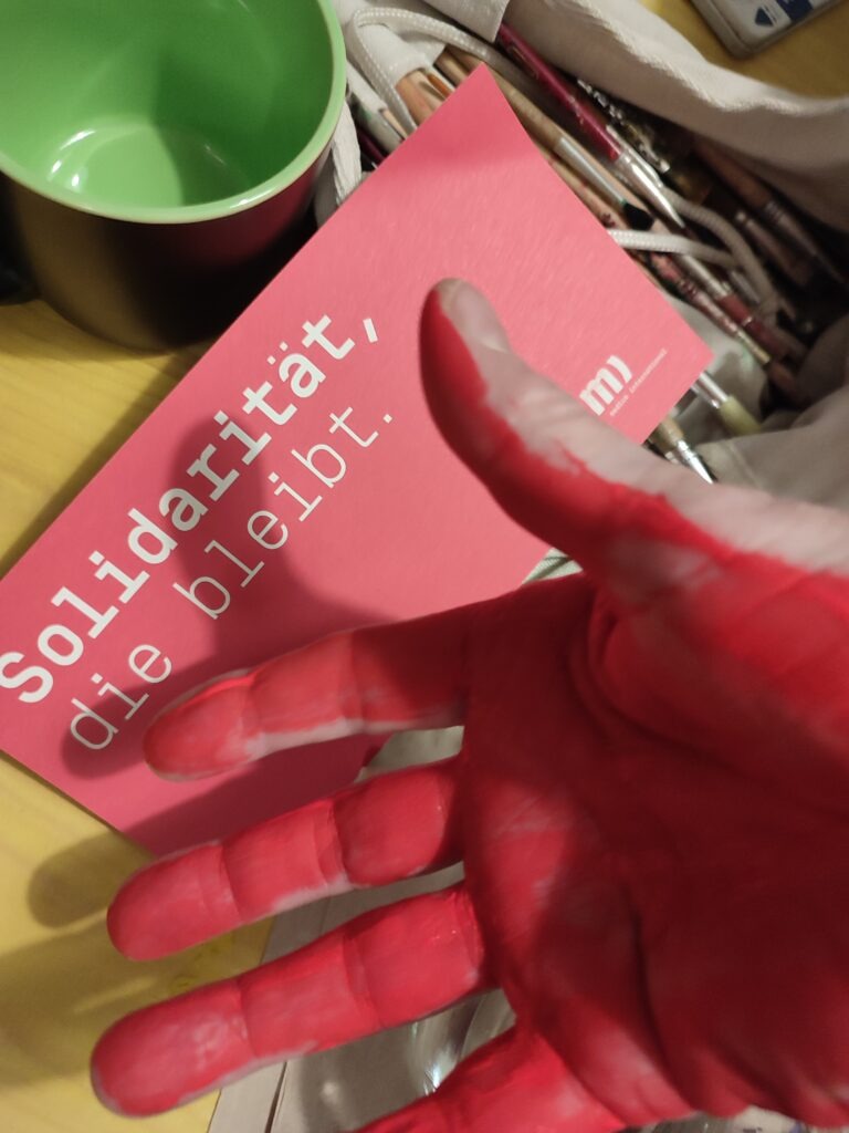 Im Vordergrund ist eine mit Farbe bemalte rote Hand. Dahinter ist eine Karte auf der steht: "Solidarität, die bleibt." Dahinter befinden sich Pinsel und eine Tasse.