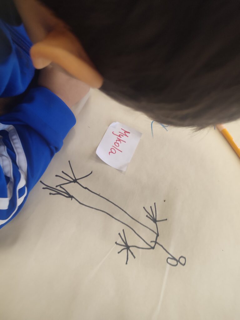 Ein Kind ist über ein Blatt Papier gebeugt, es ist nur von hinten zu sehen. Auf dem Bild ist eine Strichfigur. 
