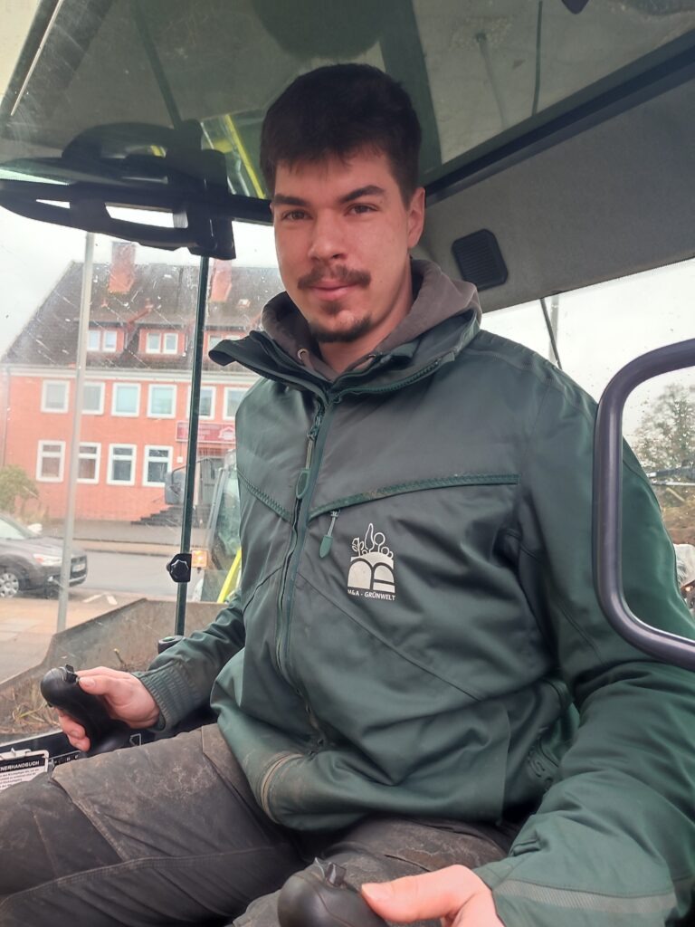 Ein Baggerfahrer sitzt in einer Fahrerkabine und schaut freundlich in die Kamera. 