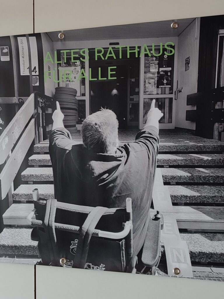 Eine Person im Rolli möchte in das Alte Rathaus. Die Auffahrhilfe fehlt jedoch (noch). Die Person zeigt auf das Gebäude. Auf dem Bild steht in grüner Schrift "Altes Rathaus für Alle".