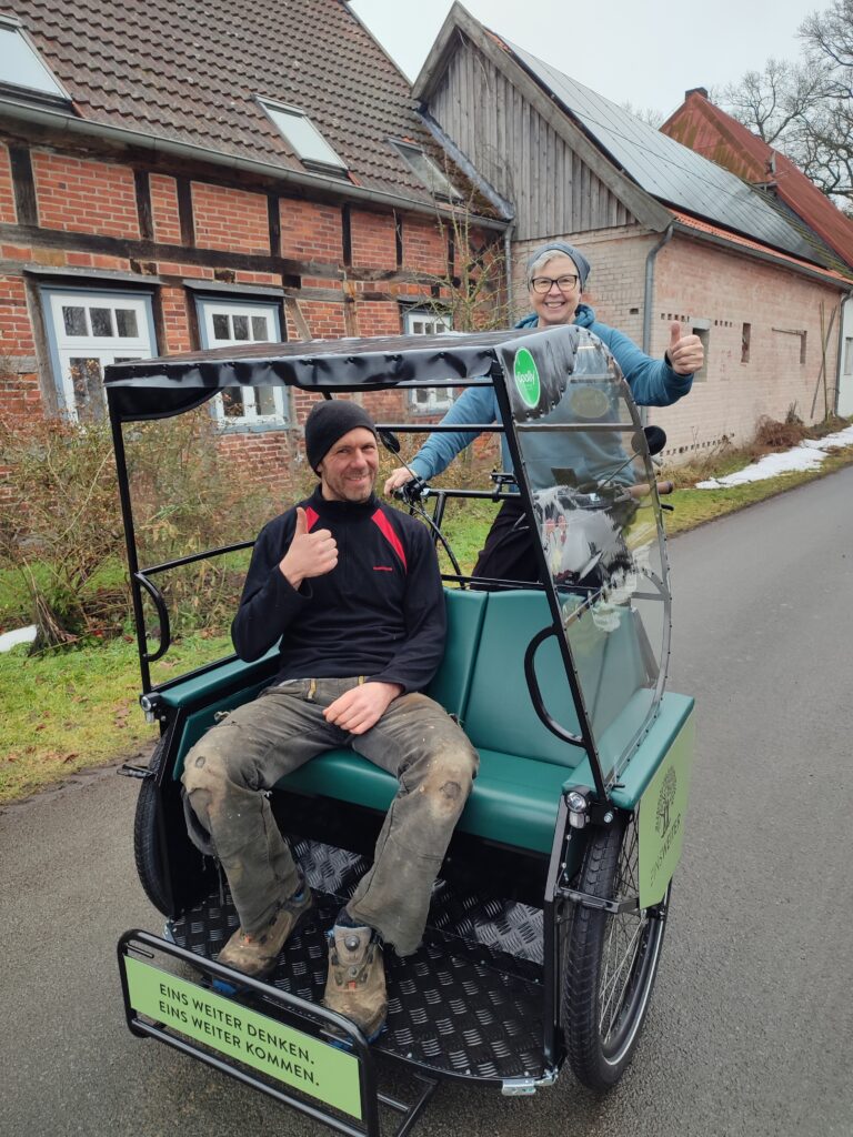 Das Foto zeigt die Fahrradrikscha. Vorne im Sitzbereich sitzt eine Person mit einer schwarzen Mütze und Arbeitskleidung und hält den Daumen hoch. Die Fahrradrikscha wird von einer Person gefahren, die ebenfalls den Daumen hoch hält. Die Rikscha steht auf einer Straße, im Hintergrund sind Fachwerkhäuser zu sehen.
