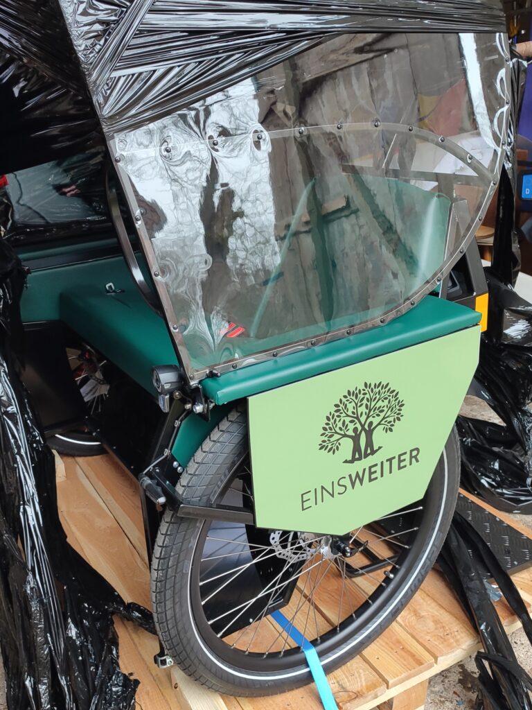 Auf dem Foto ist die Seitenansicht der Fahrradrikscha abgebildet. Auf einer grün unterlegten Fläche ist das Logo von EinsWeiter aufgedruckt.