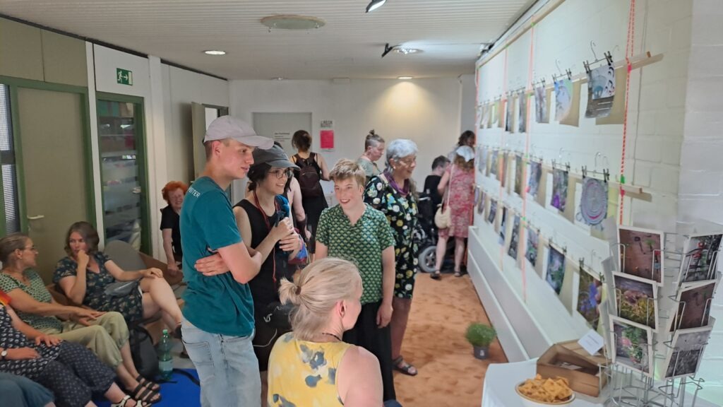 Das Foto zeigt eine Gruppe von Menschen in einem hellen Ausstellungsraum mit weißen Wänden und hellem Boden. An der rechten Wand hängen zahlreiche Fotos und Kunstwerke, die mit Wäscheklammern an einer gespannten Leine befestigt sind. Die meisten Personen stehen und betrachten die ausgestellten Bilder, einige sitzen auf Stühlen. Die Kleidung der Anwesenden ist bunt und vielfältig – mit Blumen- und geometrischen Mustern. Im Vordergrund steht ein Tisch mit Snacks. An der linken Wand ist eine grüne Tür mit Glasfenster zu erkennen, darüber ein Notausgangsschild. Die Stimmung wirkt entspannt und gesellig.
