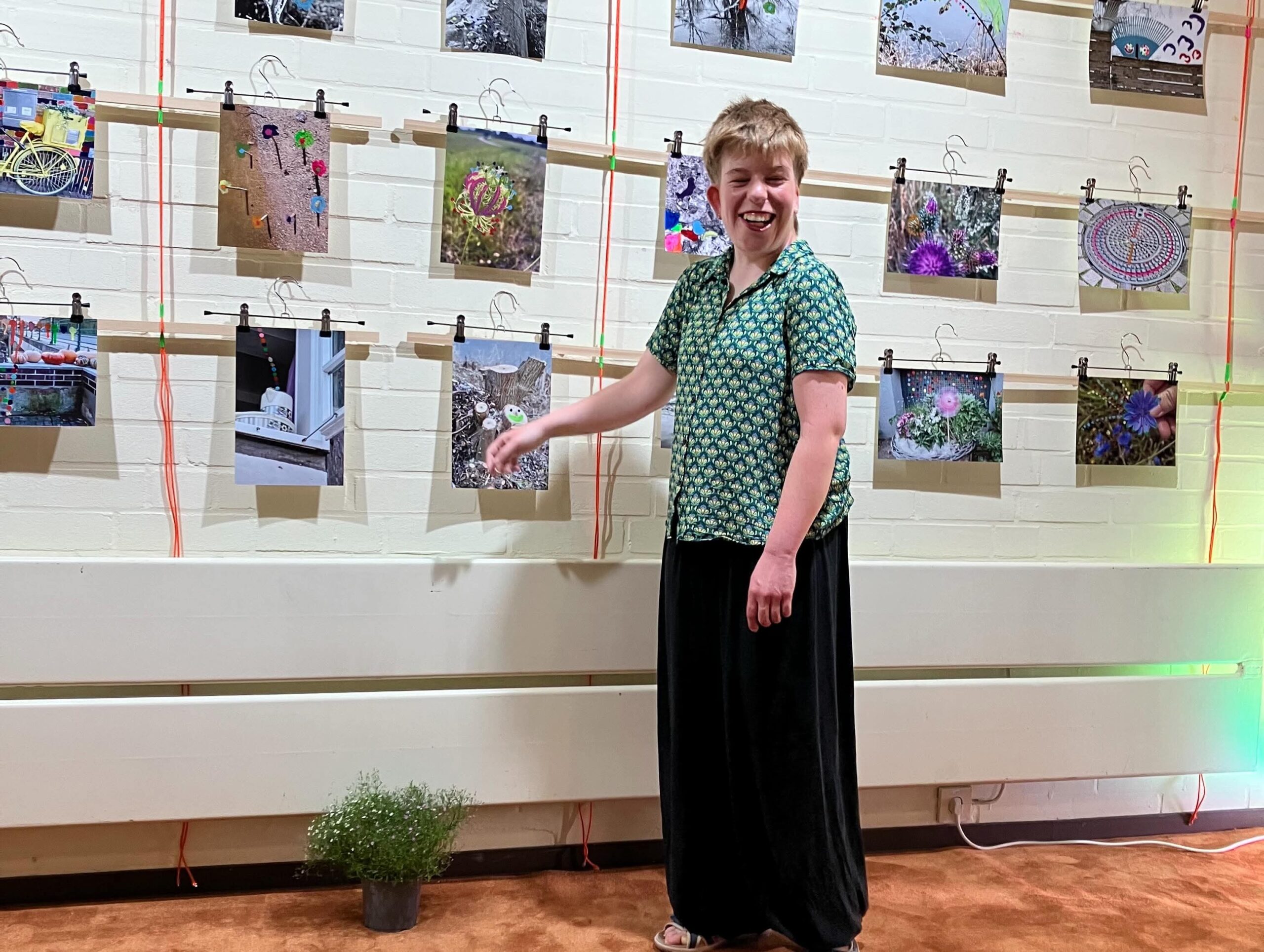 Das Bild zeigt die Künstlerin der Fotoausstellung vor einer weißen Wand, an der zahlreiche Fotos mit Wäscheklammern aufgehängt sind. Die Person trägt ein grünes, gemustertes Oberteil und einen langen, schwarzen Rock. Sie lacht und streckt den rechten Arm aus, als würde sie gerade auf eines der Fotos zeigen. Die ausgestellten Fotos zeigen verschiedene Motive – Natur, Tiere und Gegenstände –, teils farbig, teils schwarz-weiß. Zwischen den Fotos hängen bunte Fäden. Am Boden steht ein Topf mit einer kleinen Pflanze, der Boden ist ein orange-brauner Teppichboden. Die Stimmung wirkt fröhlich und lebendig.